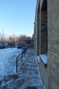A ramp with a hand rail leading to a light-coloured brick building from a parking lot.