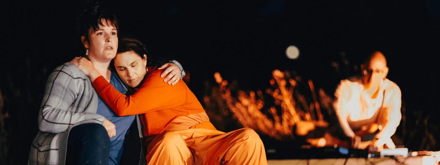 A photo from an outdoor theatre production at night. In the forefront, two women sit holding one another; in the background, a man is playing an electrical instrument, bathed in orange light.
