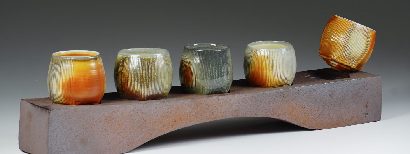 A photo of five orange, green and grey ceramic cups on a metal holder. The cup on the far right sits at an angle, appearing to balance on the edge of the holder.