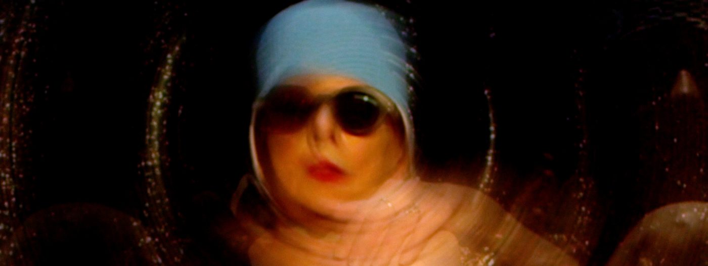 A still frame from a video of a woman wearing a blue hat and sunglasses. She is slightly out of focus.