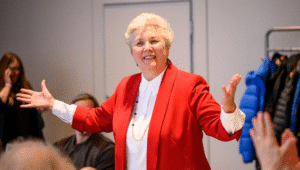 A photo of Joyce Clouston, standing in the centre of a seated audience, gesturing.