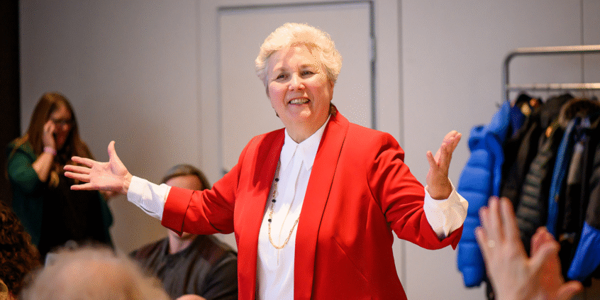 A photo of Joyce Clouston, standing in the centre of a seated audience, gesturing.