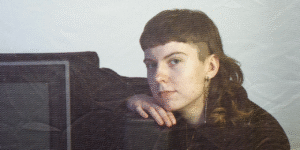 A photo of film-based artist Madelyn Gowler. They are resting their head on their land, leaning on a tube TV.