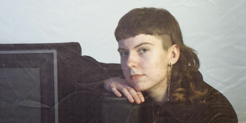 A photo of film-based artist Madelyn Gowler. They are resting their head on their land, leaning on a tube TV.