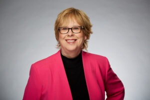 A photo of Judy Murphy. She is smiling at the camera, wearing black rectangular glasses and a magenta blazer.